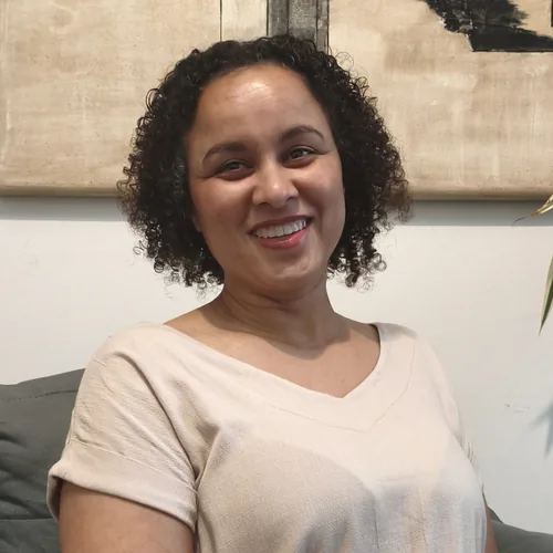 Retrato de Elaine Aparecida Correia sentada em uma poltrona cinza, sorrindo para a câmera. Ela veste uma blusa clara e, ao fundo, há uma obra abstrata em tons neutros.