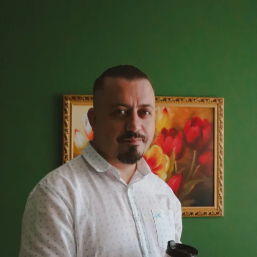 Elcio Marcio Kava, psicólogo, em pé segurando um copo térmico, usando camisa social branca, em ambiente interno com parede verde e quadro floral ao fundo.