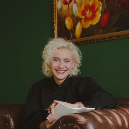 Daniela Coelho Zys, psicóloga, sentada em uma poltrona com bloco de anotações e caneta nas mãos, em ambiente interno com parede verde e quadro floral ao fundo.