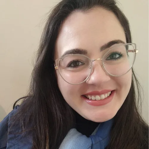 Retrato de Adriane Silva de Azevedo sorrindo para a câmera. Ela usa óculos de armação transparente, casaco azul e um cachecol em tons de azul, em um ambiente interno com fundo claro.